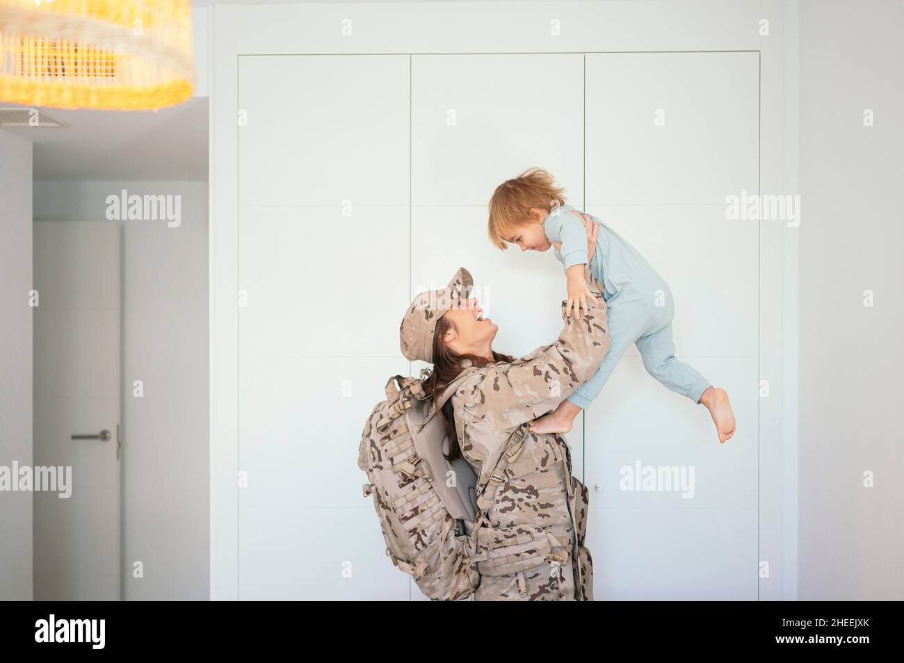 Vista laterale di felice soldato femminile in uniforme con zaino che tiene il figlio carino in braccia distese vicino al presepe in legno in camera da letto Foto Stock