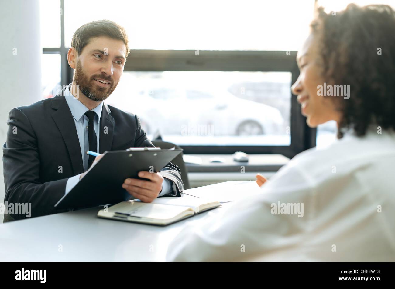 Intervista, concetto di assunzione. Caucasica successo CEO società, assumere manager, intervistando una donna afroamericana candidato, esaminando il suo curriculum, ascoltando le informazioni, in ufficio moderno, sorride Foto Stock