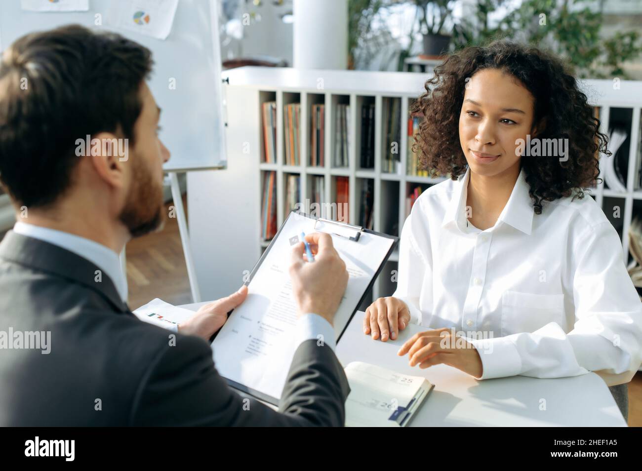 Una giovane donna afroamericana intelligente in abiti formali, viene a ottenere un lavoro in un'azienda, viene intervistato con il suo capo, mostra il suo curriculum, sorridente. Il responsabile di assunzione che guarda il resume, conduce l'intervista Foto Stock