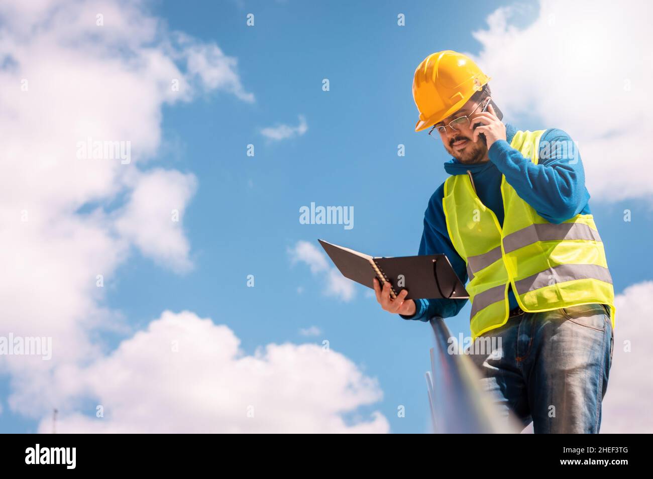 lavoratore latino con elmetto giallo, occhiali e gilet verde, appoggiato su una ringhiera con un diario in mano, parlando al telefono. Foto Stock