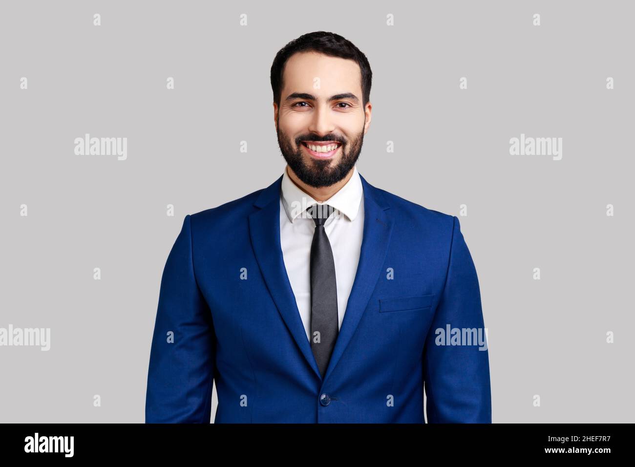 Uomo allegro con sorriso affascinante, in piedi e guardando la macchina fotografica, esprimendo emozioni positive, indossando un abito di stile ufficiale. Studio interno girato isolato su sfondo grigio. Foto Stock