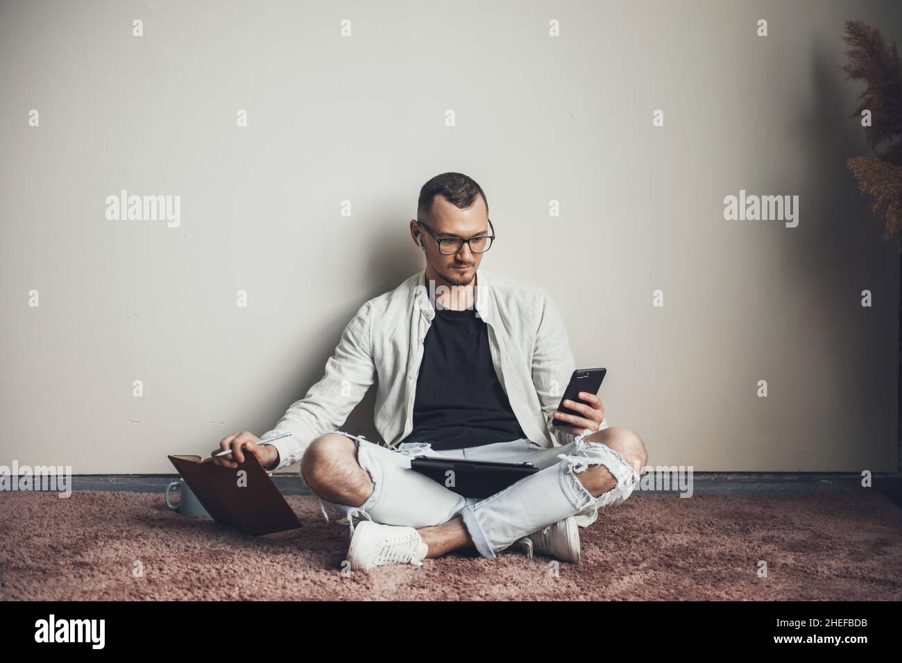 Vista frontale di un uomo caucasico seduto sul pavimento guardando lo smartphone mentre tiene un libro. Comunicazione mobile. Formazione online. Foto Stock