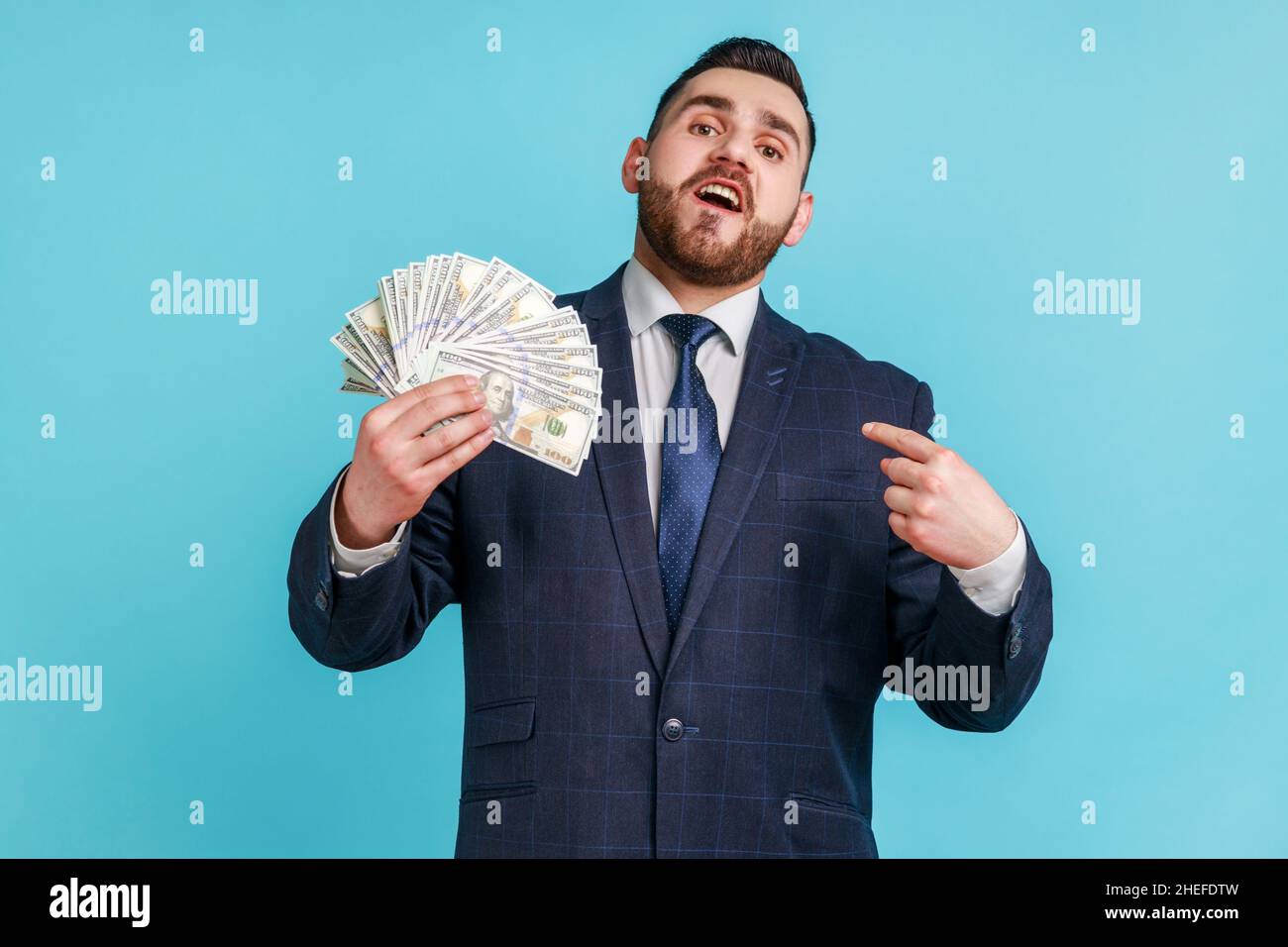 Ricco uomo d'affari portatore di vestito scuro di stile ufficiale, puntando il dito su se stesso e mostrando molte banconote in dollari, orgoglioso della sua ricchezza e successo. Studio interno girato isolato su sfondo blu Foto Stock