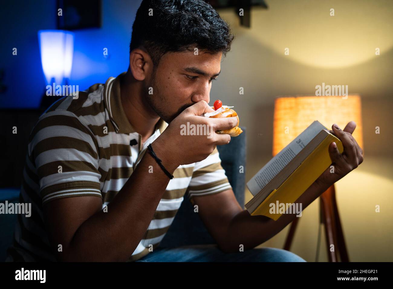 Giovane studente indiano che mangia cibo malsano mentre legge il libro a casa - concetto di stile di vita malsano, hobby, abilità e sviluppo di knowladge Foto Stock