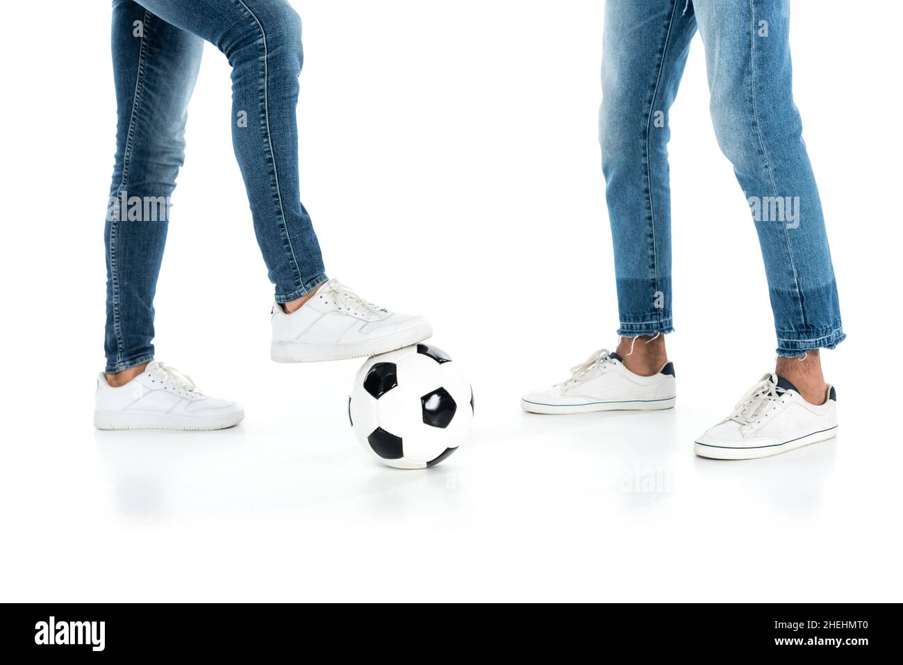 vista ritagliata di uomini interrazziali in jeans e sneakers che giocano a calcio su bianco Foto Stock