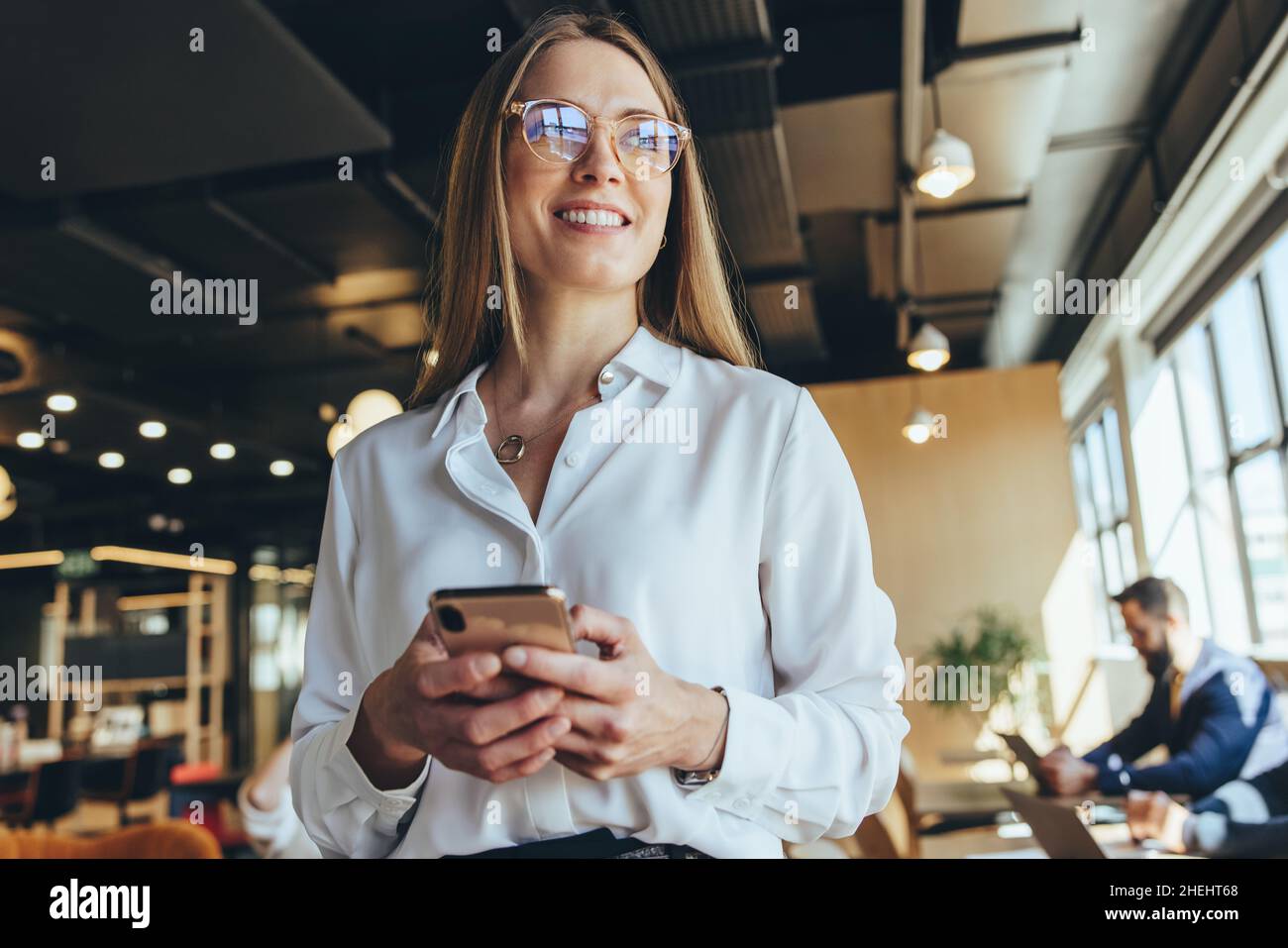 Donna d'affari premurosa che tiene uno smartphone in uno spazio di co-lavoro. Felice giovane donna d'affari che guarda via con un sorriso mentre si levano in piedi in un wo moderno Foto Stock