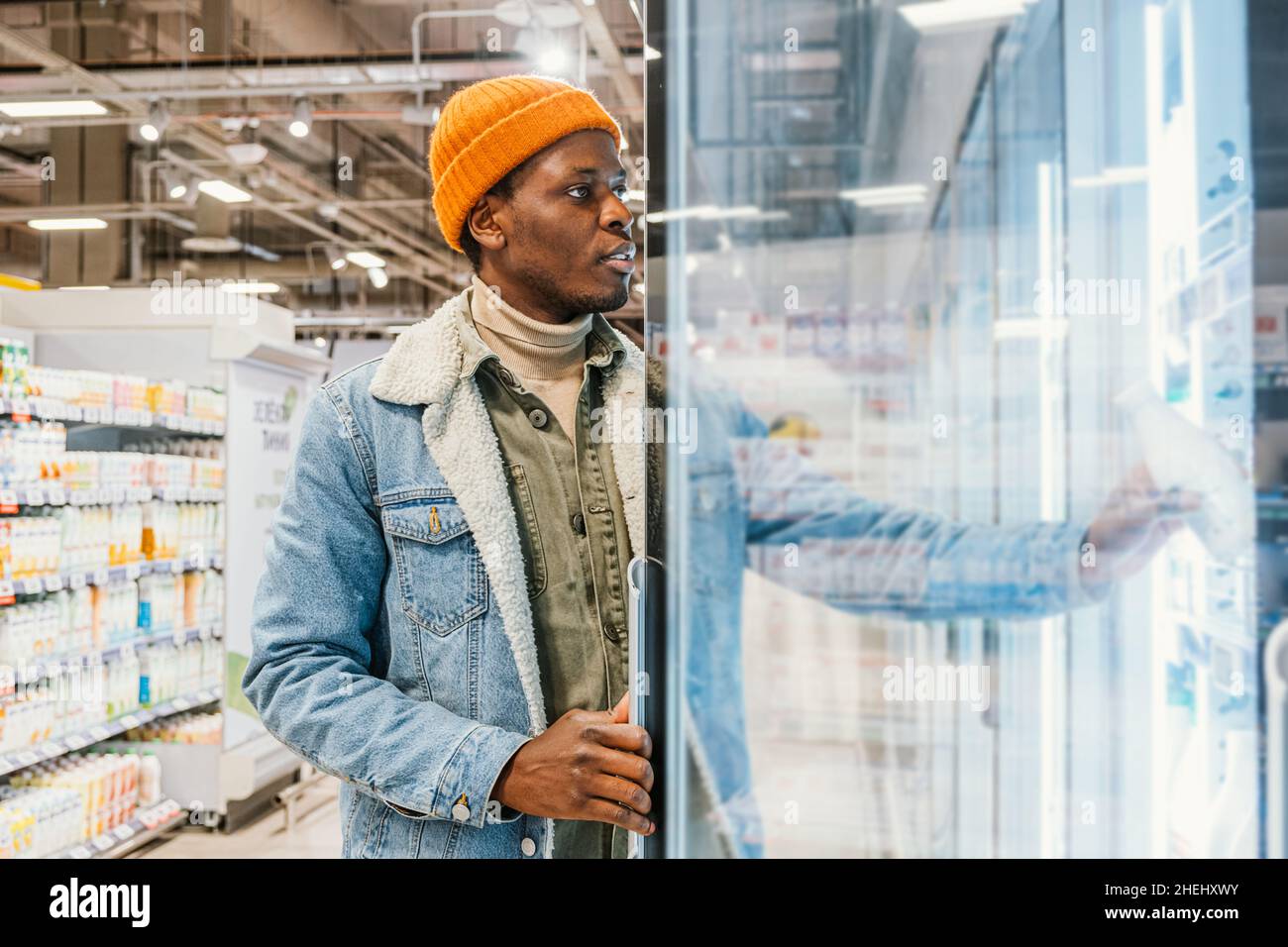 Il ragazzo afro-americano positivo in giacca di denim e cappello arancione prende la bottiglia di latte dalla scatola del frigorifero in vista laterale del moderno supermercato Foto Stock