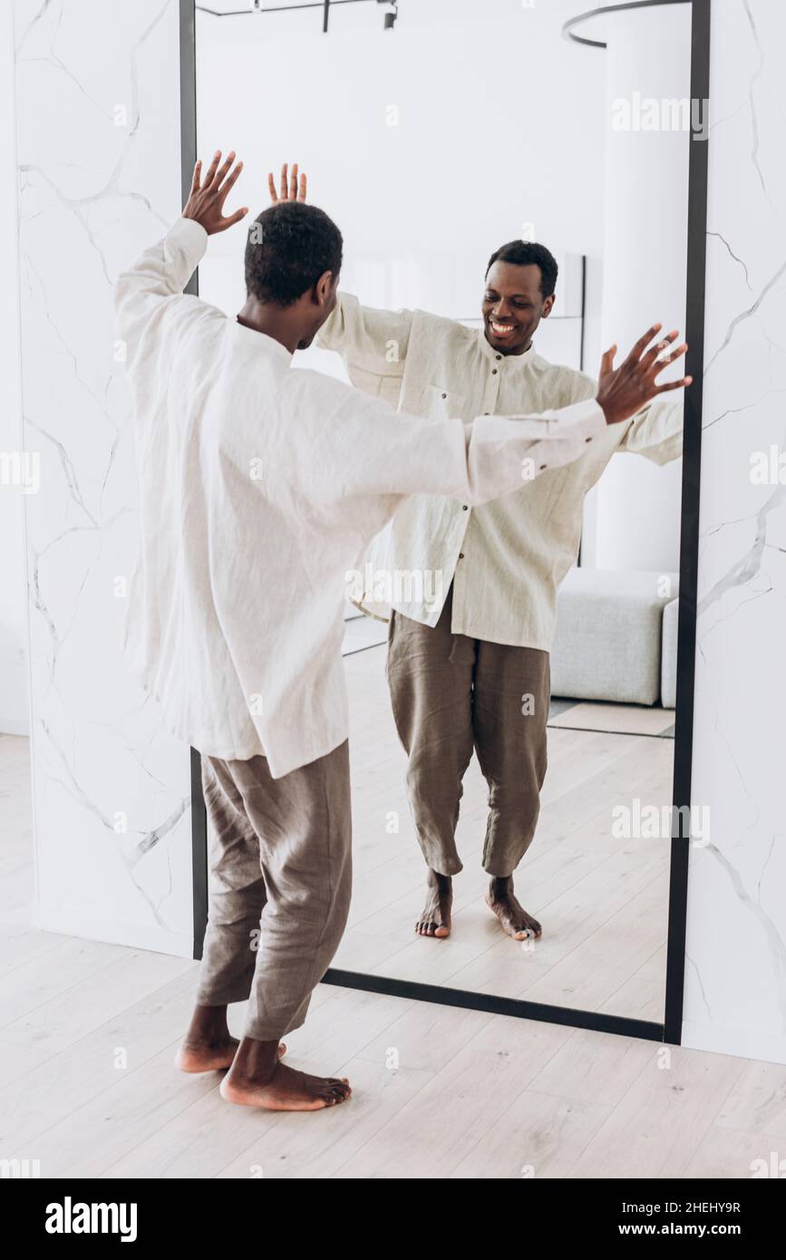 Positivo giovane africano-americano ragazzo in sciolto camicia bianca e pantaloni danze guardando grande specchio riflessione a casa dietro la vista Foto Stock