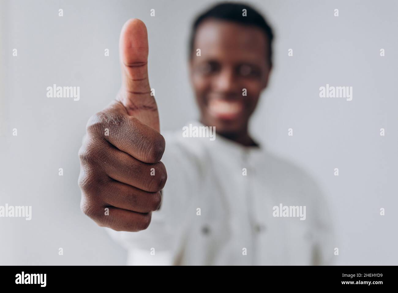 Happy afroamericano blurry persona in camicia bianca mostra pollice in posa per la fotocamera su sfondo grigio chiaro closeup estremo Foto Stock