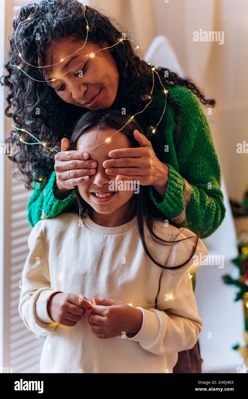 Bruna scolgirl e curly afroamericana madre posare gli occhi chiudendo con le mani in garland illuminante e sorridere in elegante soggiorno Foto Stock