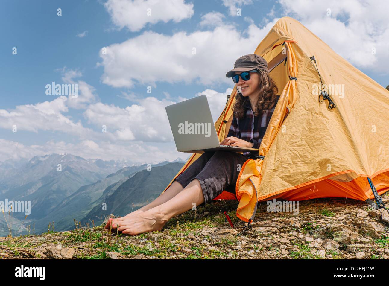 Una donna freelance felice sta lavorando ad un laptop in una tenda arancione alta nelle montagne in una giornata estiva soleggiata contro un cielo blu Foto Stock