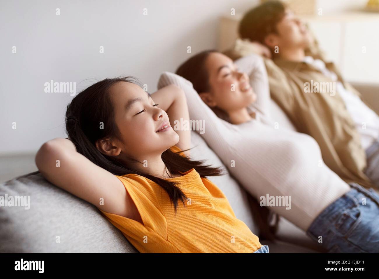 Ragazza asiatica soddisfatta, mamma millenaria e papà rilassarsi sul divano in silenzio in camera interna minimalista a tempo libero Foto Stock