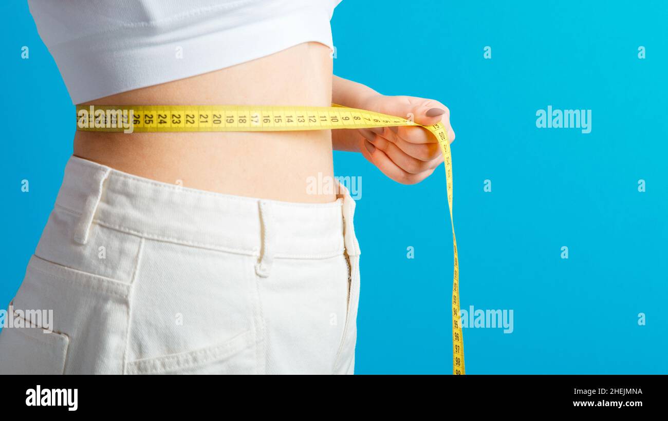 Donna sottile misura la sua linea vita con nastro di misurazione. Corpo sano che forma il concetto di perdita del peso. Vita sottile piccolo ventre in grandi pantaloni jeans bianchi Foto Stock