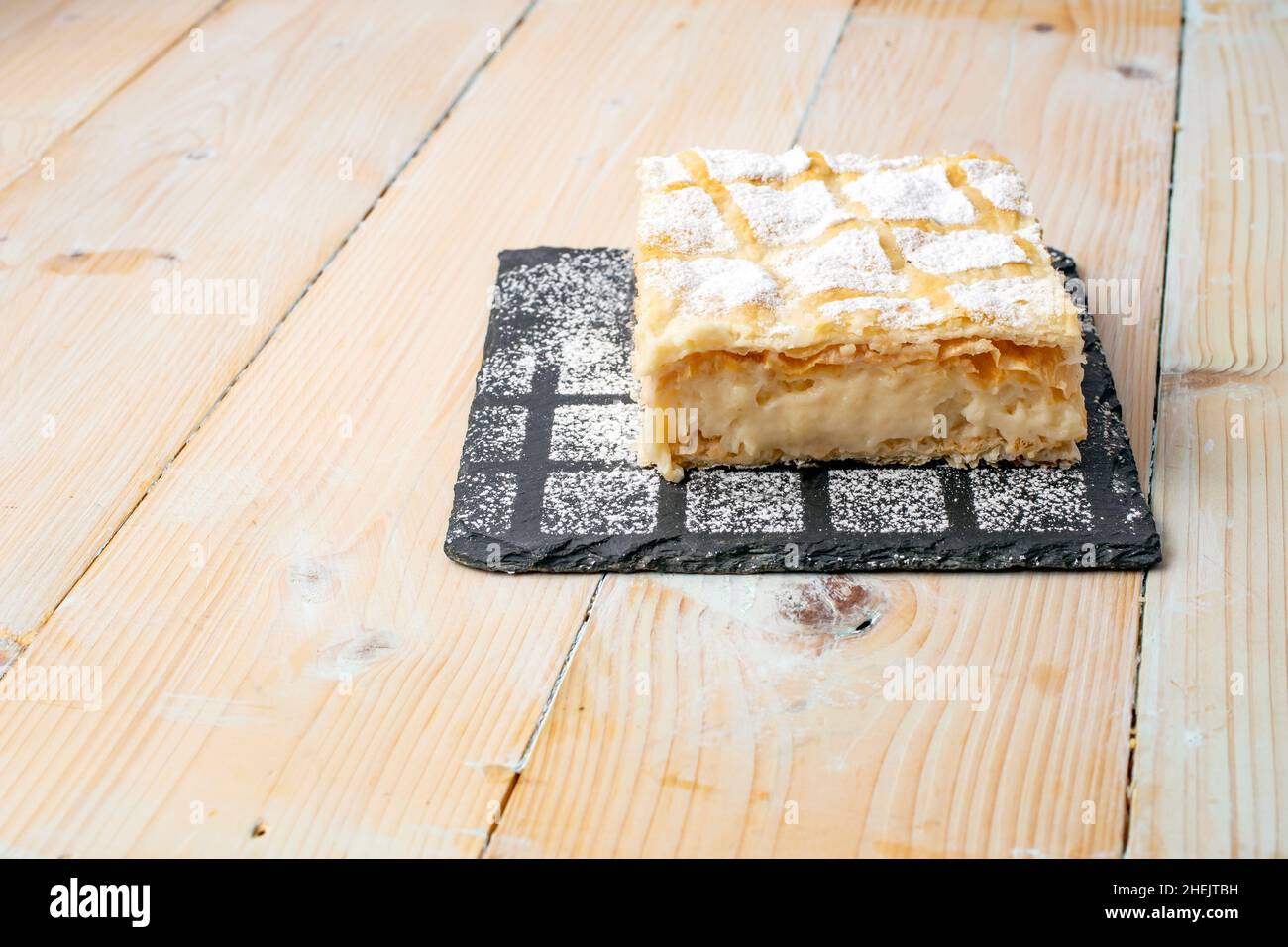 cremeschnitte o cremsnit torta su una pietra quadrata nera mattonella trama con polvere di zucchero cosparso e un bianco lavati tavole di legno tavola Foto Stock