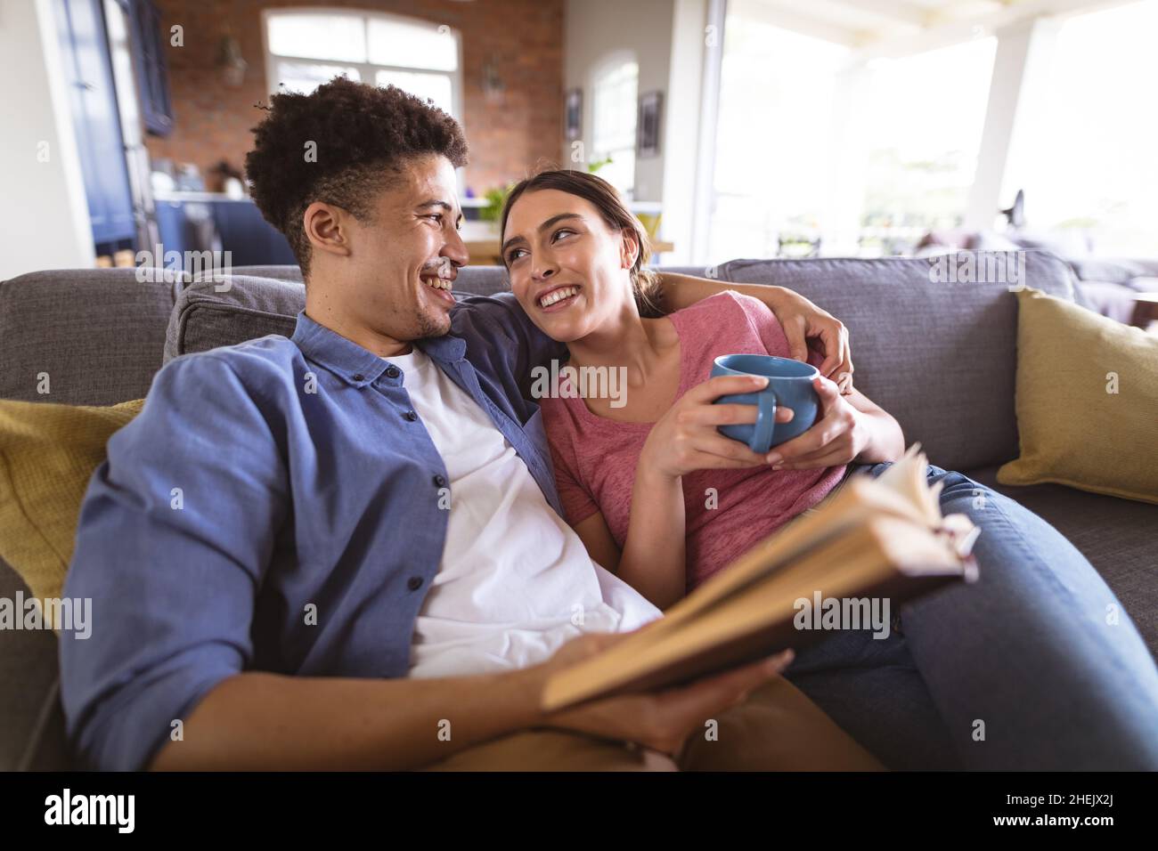 Felice coppia biraciale amorevole che si guarda l'un l'altro mentre si rilassa con caffè e libro a casa Foto Stock