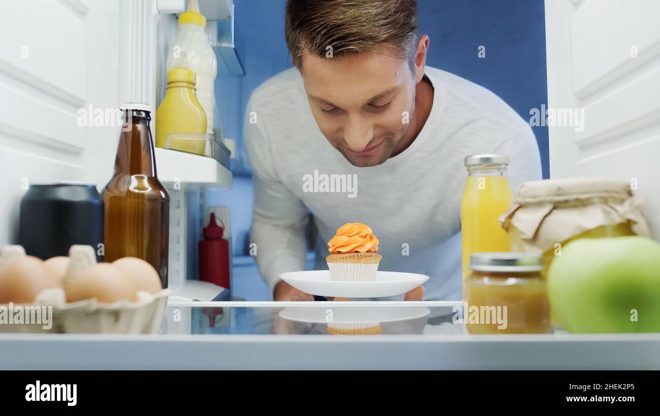 uomo sorridente che prende cupcake dal frigorifero vicino a bevande, uova, vasetti e contenitori con cibo Foto Stock