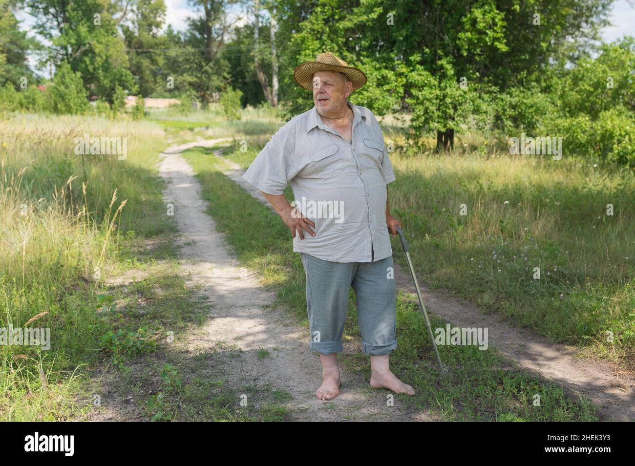 Ritratto di contadino maggiore ucraino in cappello in piedi su una strada di campagna a piedi nudi nella stagione estiva Foto Stock
