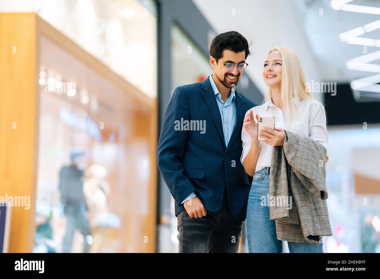 Allegro bella coppia giovane utilizzando il telefono cellulare in piedi nella hall del centro commerciale. Uomo bearded in vetri e donna bionda che compra al centro del deposito. Foto Stock
