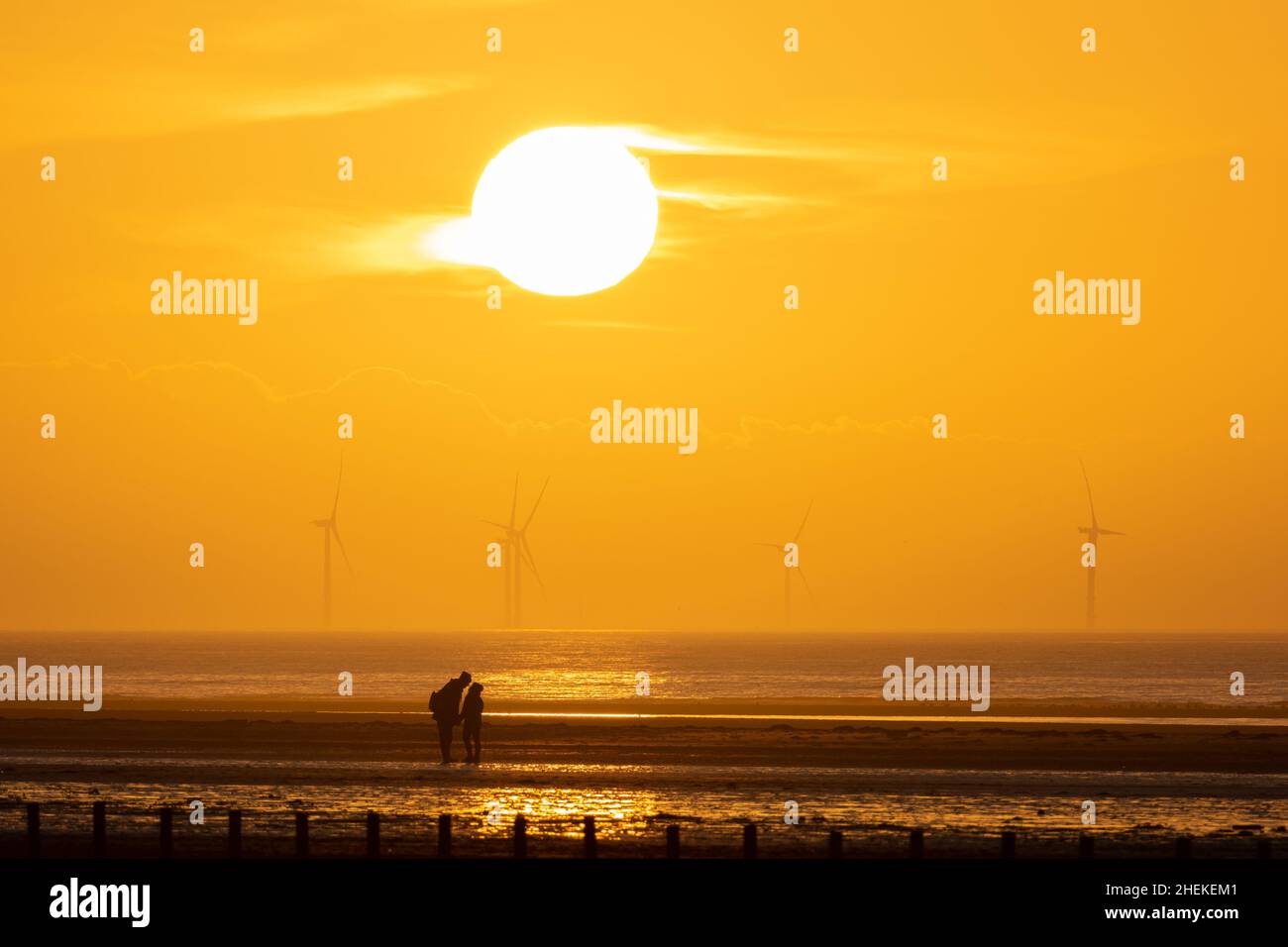 Ainsdale Beach, Southport, Merseyside, Regno Unito. 11th Jan 2022. Il sole si prepara a tramontare dietro turbine eoliche lontane mentre il cielo si allontano, con l'alta pressione che si muove e le condizioni nel Nord Ovest che saranno sistemate per tutta la settimana. Una coppia si erge sotto il sole. Credit: Callum Fraser/Alamy Live News Foto Stock