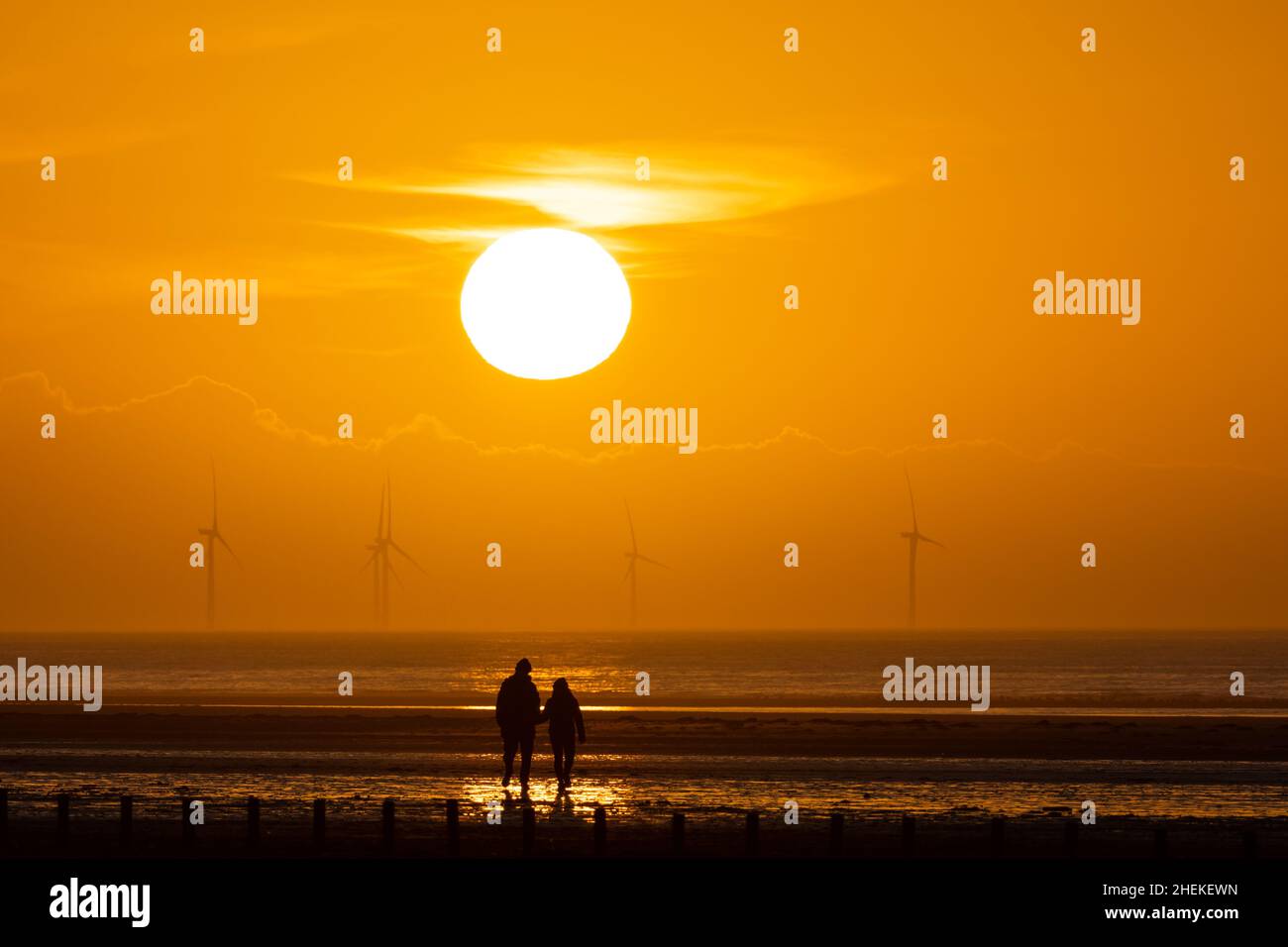 Ainsdale Beach, Southport, Merseyside, Regno Unito. 11th Jan 2022. Il sole si prepara a tramontare dietro turbine eoliche lontane mentre il cielo si allontano, con l'alta pressione che si muove e le condizioni nel Nord Ovest che saranno sistemate per tutta la settimana. Una coppia si erge sotto il sole. Credit: Callum Fraser/Alamy Live News Foto Stock