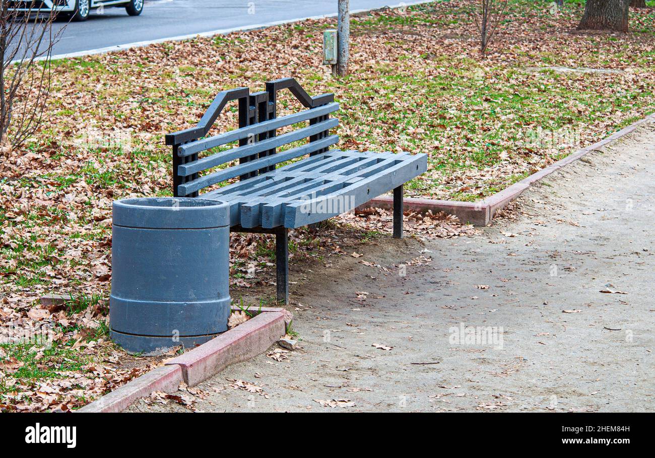 Una panca di legno dipinta di grigio e un cestino in un parco autunnale vicino al sentiero. Foto Stock