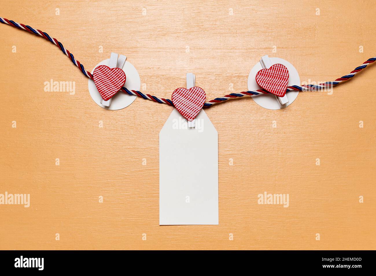 Minimalistico semplice concetto di San Valentino, carta bianca vuota per nomi o desideri con piccoli perni di cuore rosso su sfondo di legno beige Foto Stock
