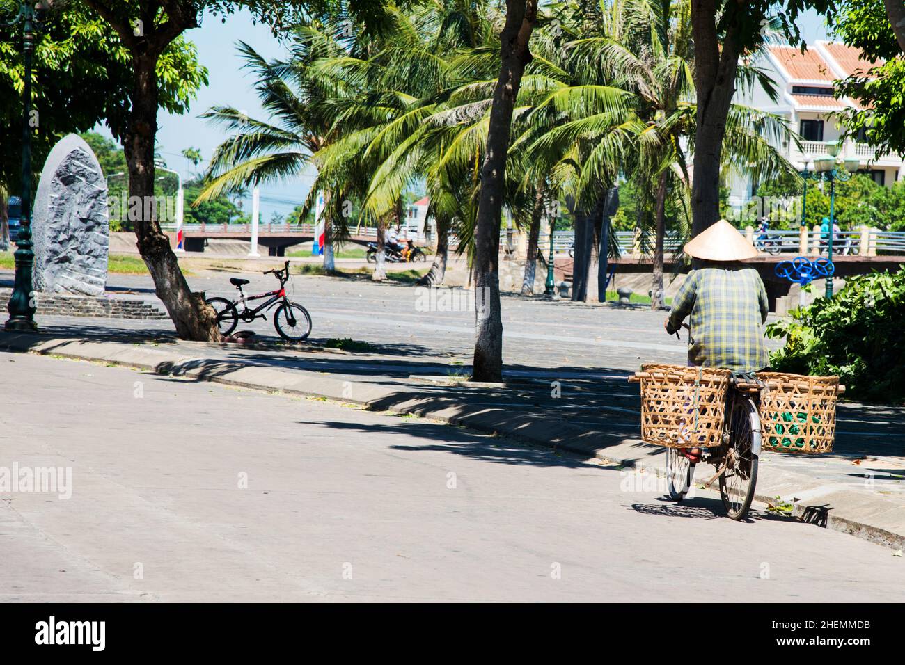 Persona vista dalla schiena muovendosi su una bicicletta con un cappello conico e un palo da trasporto con verdure. Strada vuota. Hue, Vietnam, Asia Foto Stock