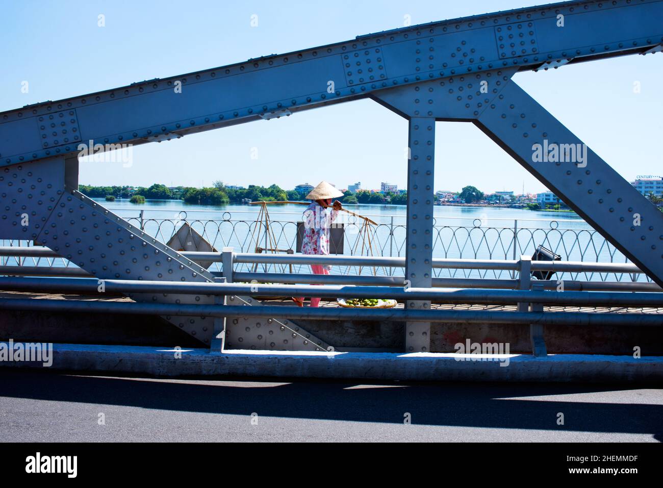 Persona irriconoscibile che va al mercato con un cappello di paglia conico e un palo da trasporto. Attraversando il ponte Hue. Vietnam. Asia Foto Stock