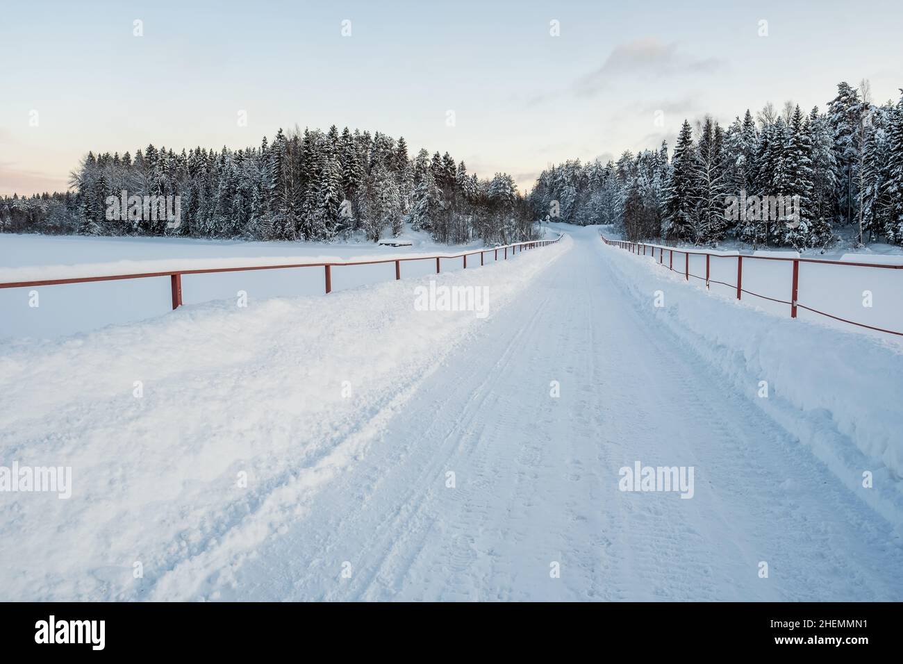 Ponte rurale con una strada sgevera dalla neve sullo sfondo di un grande lago ghiacciato, foresta e cielo. Bellissimo paesaggio invernale. Foto Stock