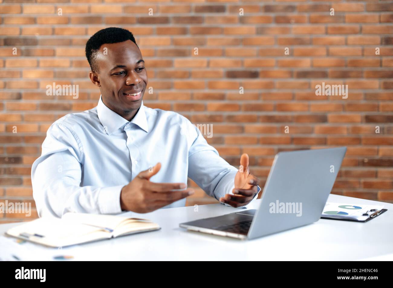 Conversazione remota. L'uomo d'affari afro-americano di successo, l'azienda del ceo, il responsabile di vendite, usa il laptop per la comunicazione a distanza con il cliente, siede nell'ufficio moderno, gesturing con le sue mani, sorridente Foto Stock