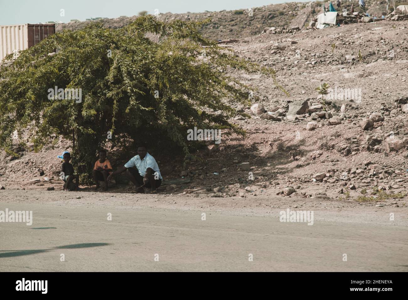 Gibuti, Gibuti - 21 maggio 2021: Un Gibutiano e i suoi due figli seduti sulla pietra e in attesa dell'autobus all'ombra di un albero a Gibuti. Editor Foto Stock