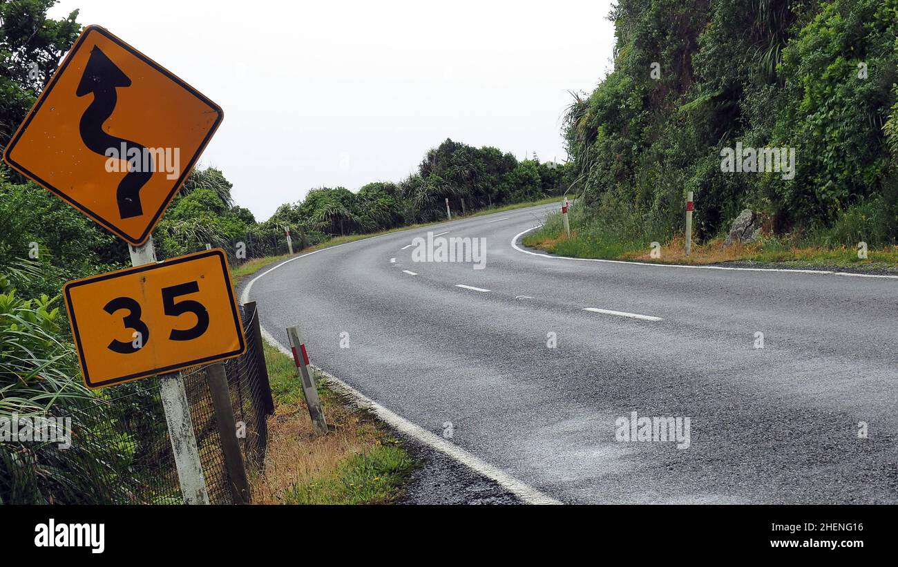 Un angolo stretto e tortuosa strada che arriva su una costa occidentale NZ strada. il segno 35kph è la velocità consigliata per affrontare le curve in modo sicuro Foto Stock