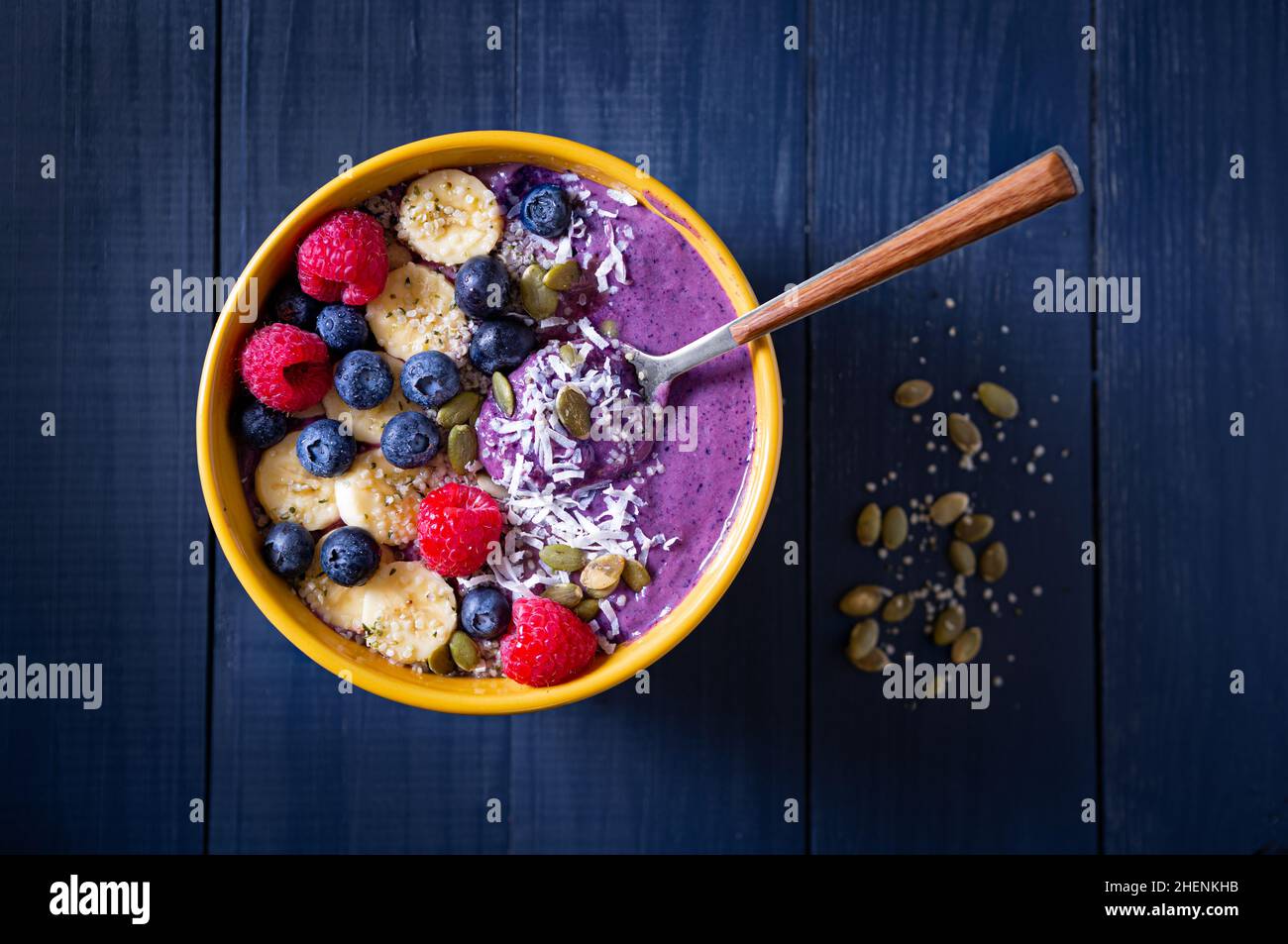 Ciotola sana di frullato di mirtillo con banana, lampone, semi di zucca, semi di canapa e cocco in una ciotola gialla su una superficie di legno blu Foto Stock