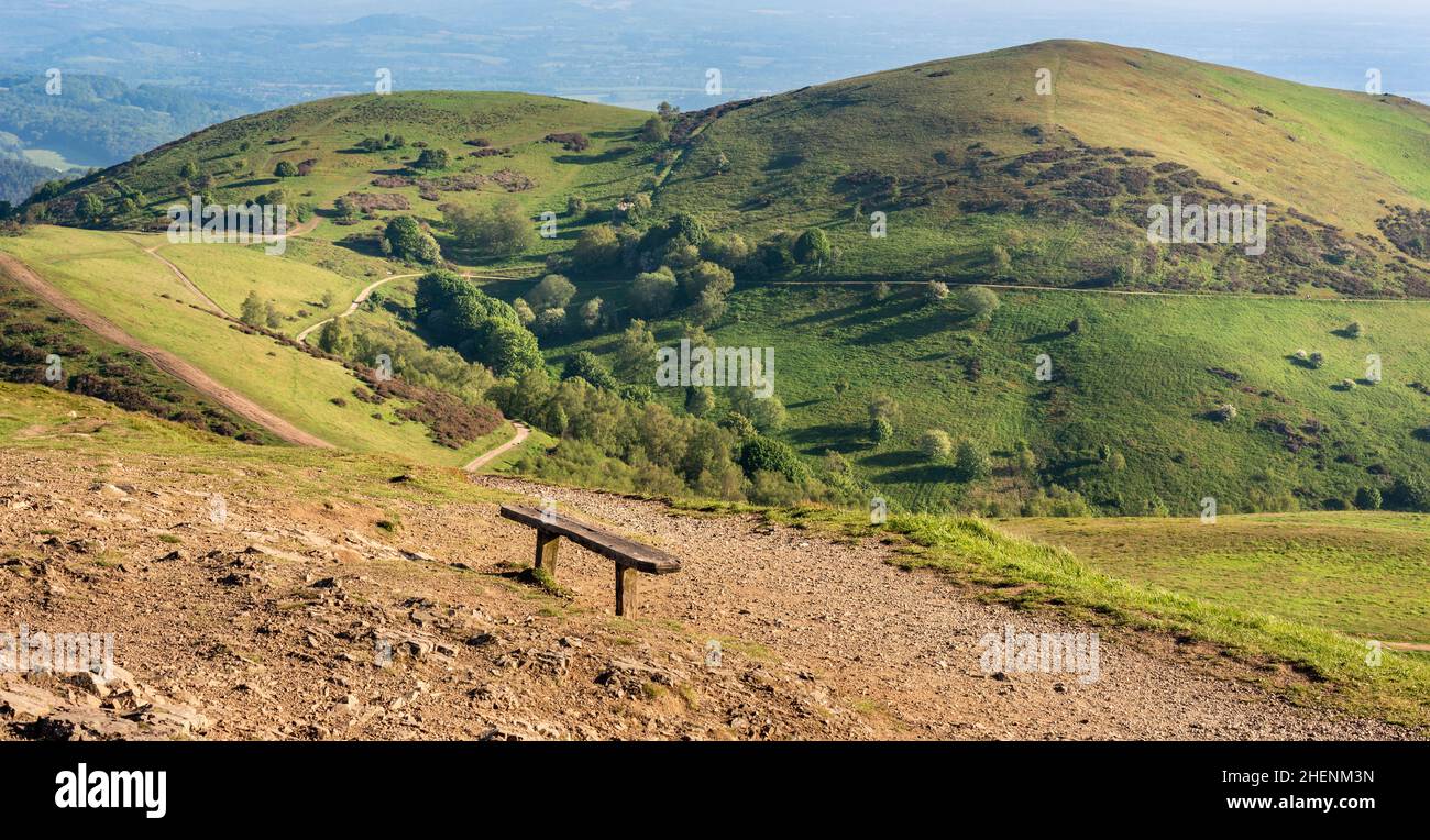 Una panca di legno e punto di vista dalla cima più alta di Malvern, guardando verso Table Hill e North Hill coperto di verde lussureggiante erba, all'inizio di giugno soleggiato su Foto Stock