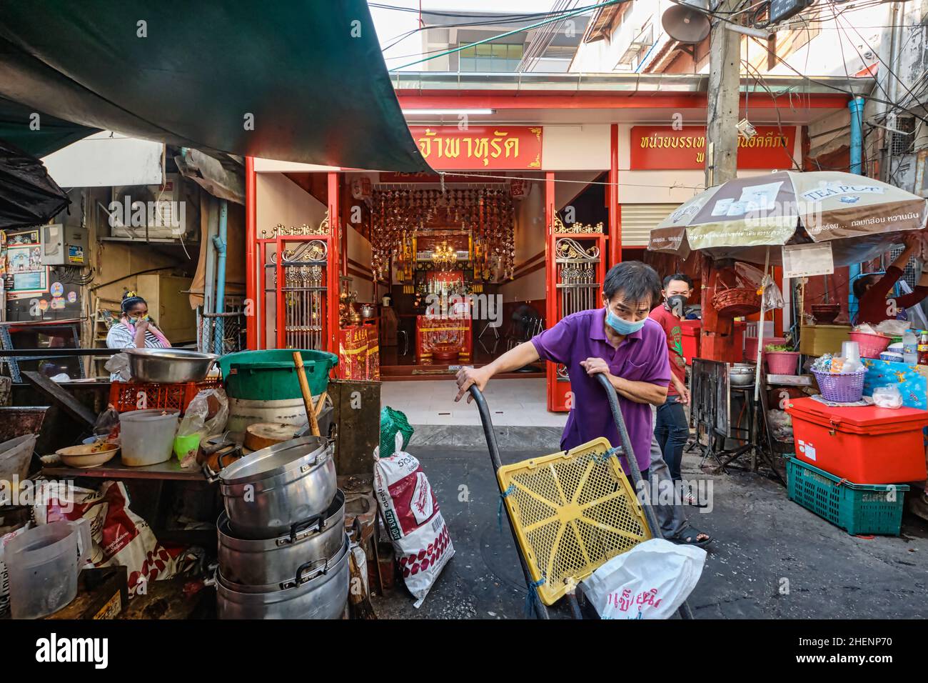 Un operaio del mercato spinge il suo carrello attraverso una corsia in Pahurat (Pharurat) mercato a Bangkok, Thailandia, un piccolo tempio cinese taoista sullo sfondo Foto Stock
