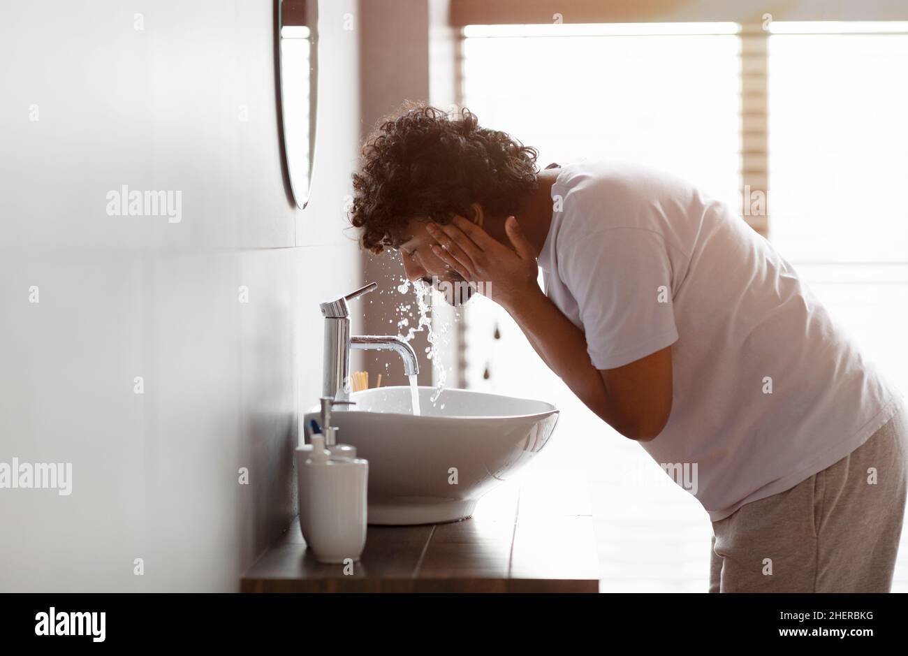 Il giovane uomo indiano che lava faccia con acqua pulita dal rubinetto che si piega sopra il lavandino, facendo la cura della pelle di routine viso in bagno Foto Stock