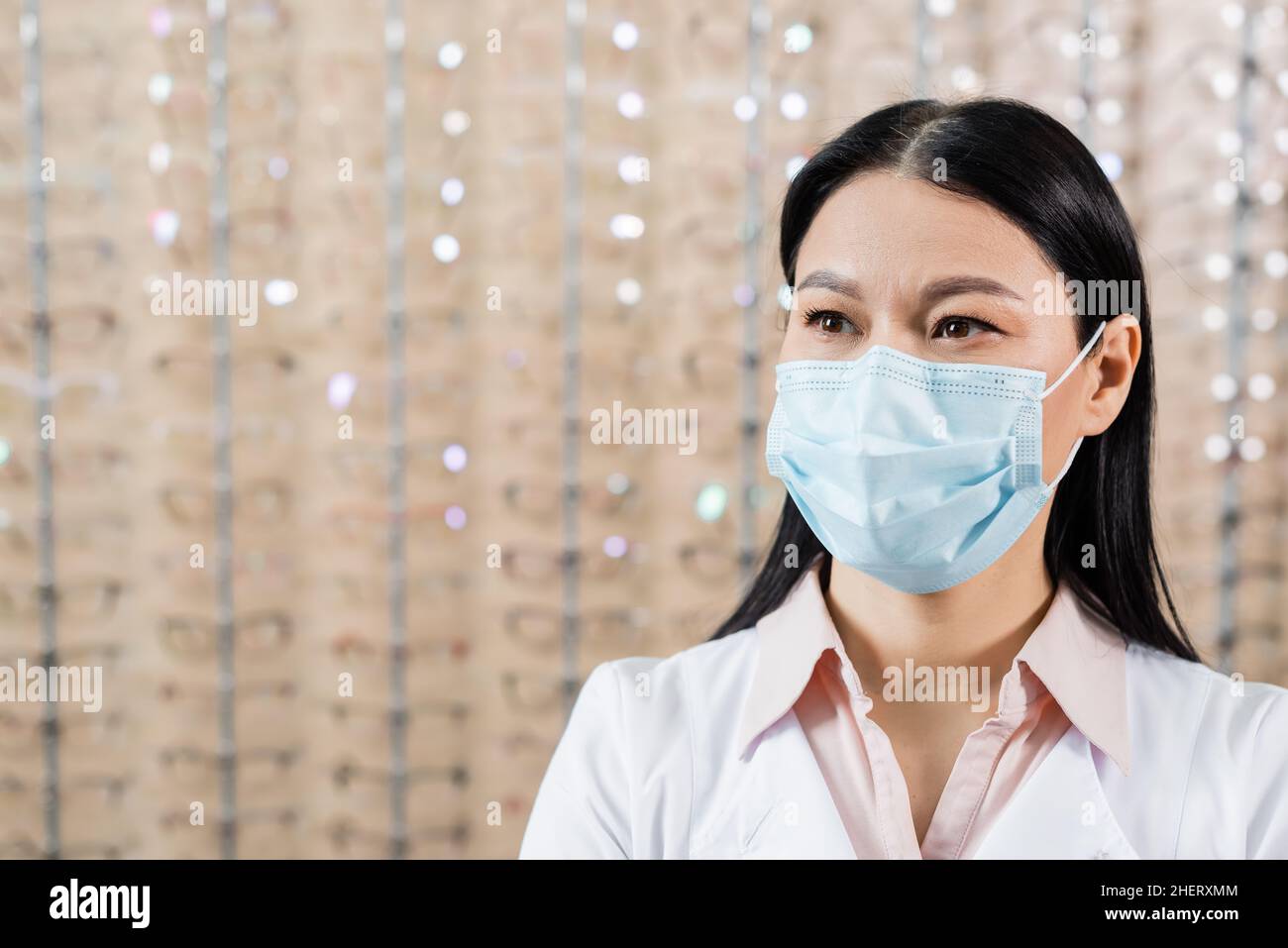 oculista asiatico in maschera medica vicino offuscato assortimento di occhiali in ottica salone Foto Stock
