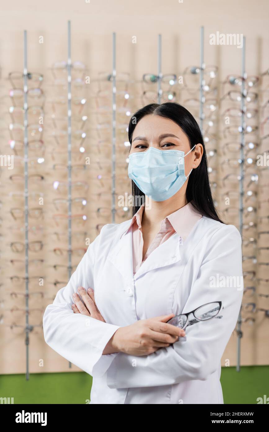 oftalmologo asiatico in maschera medica in piedi con braccia incrociate vicino offuscato assortimento di occhiali in negozio di ottica Foto Stock
