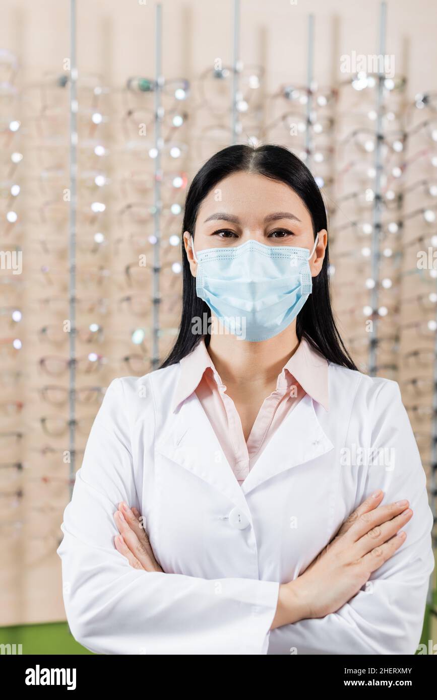 oftalmologo asiatico in maschera medica in piedi con braccia incrociate in salone di ottica Foto Stock