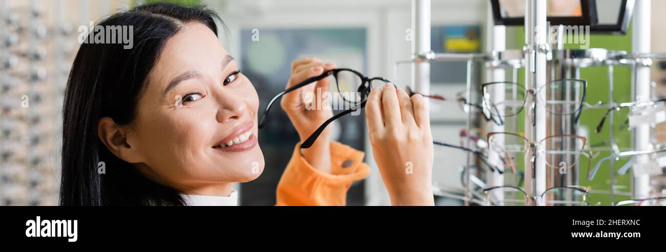 donna asiatica felice guardando la macchina fotografica mentre sceglie gli occhiali in salone di ottica, banner Foto Stock