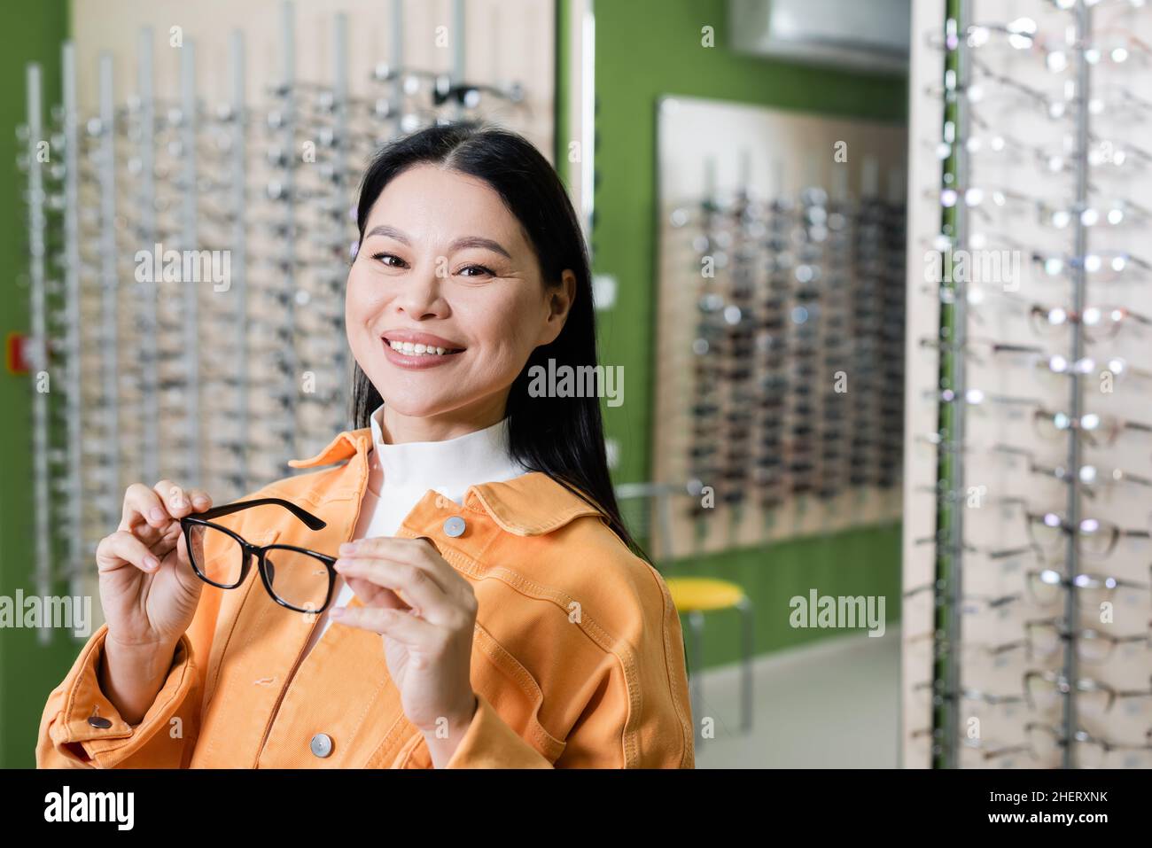 donna asiatica felice guardando la macchina fotografica mentre tiene gli occhiali in negozio di ottica Foto Stock