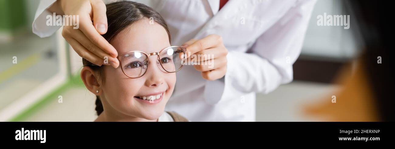 oculista cercando occhiali sulla ragazza sorridente vicino alla madre sfocata in negozio di ottica, banner Foto Stock