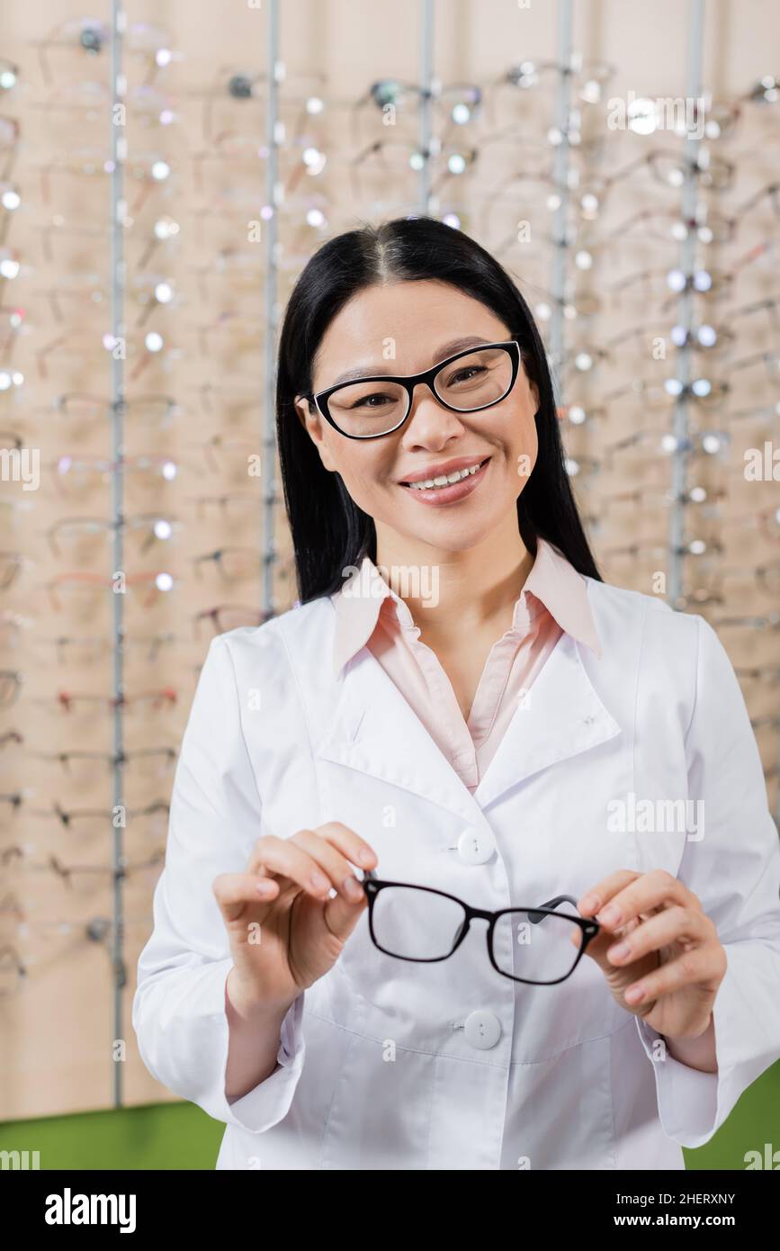 allegro oftalmologo asiatico che guarda la macchina fotografica mentre tiene gli occhiali in negozio di ottica Foto Stock