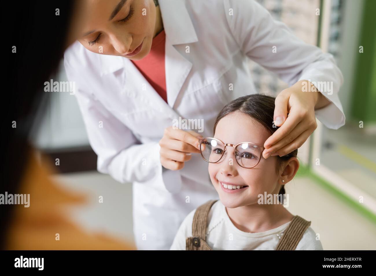 oftalmologo cercando occhiali sulla ragazza sorridente vicino mom sfocato in negozio di ottica Foto Stock