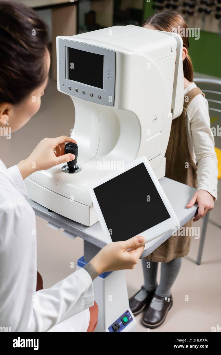 oftalmologo blurred che tiene il tablet digitale con schermo bianco mentre misura la vista della ragazza su autorefractor nel deposito dell'ottica Foto Stock
