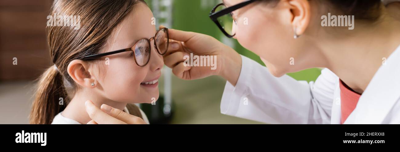 oftalmologo cercando occhiali sulla ragazza sorridente in negozio di ottica, banner Foto Stock