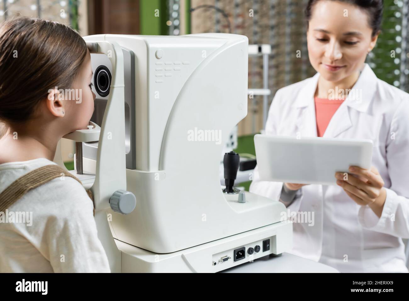 optometristo positivo guardando il tablet digitale mentre si misura la visione della ragazza su autorefrattore Foto Stock