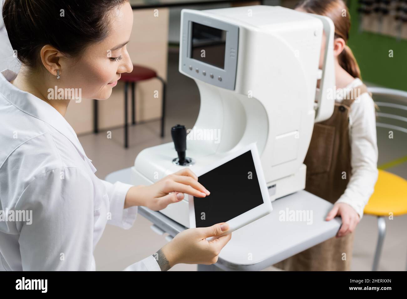 oculista sorridente utilizzando un tablet digitale con schermo vuoto mentre si misura la visione di una ragazza sfocata su oftalmoscopio Foto Stock