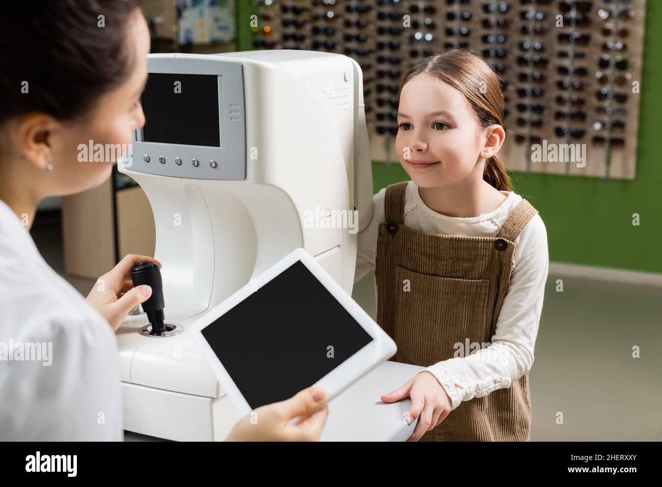 sfocato oculista che tiene tablet digitale con schermo vuoto vicino a ragazza positiva e visione screener in negozio di ottica Foto Stock