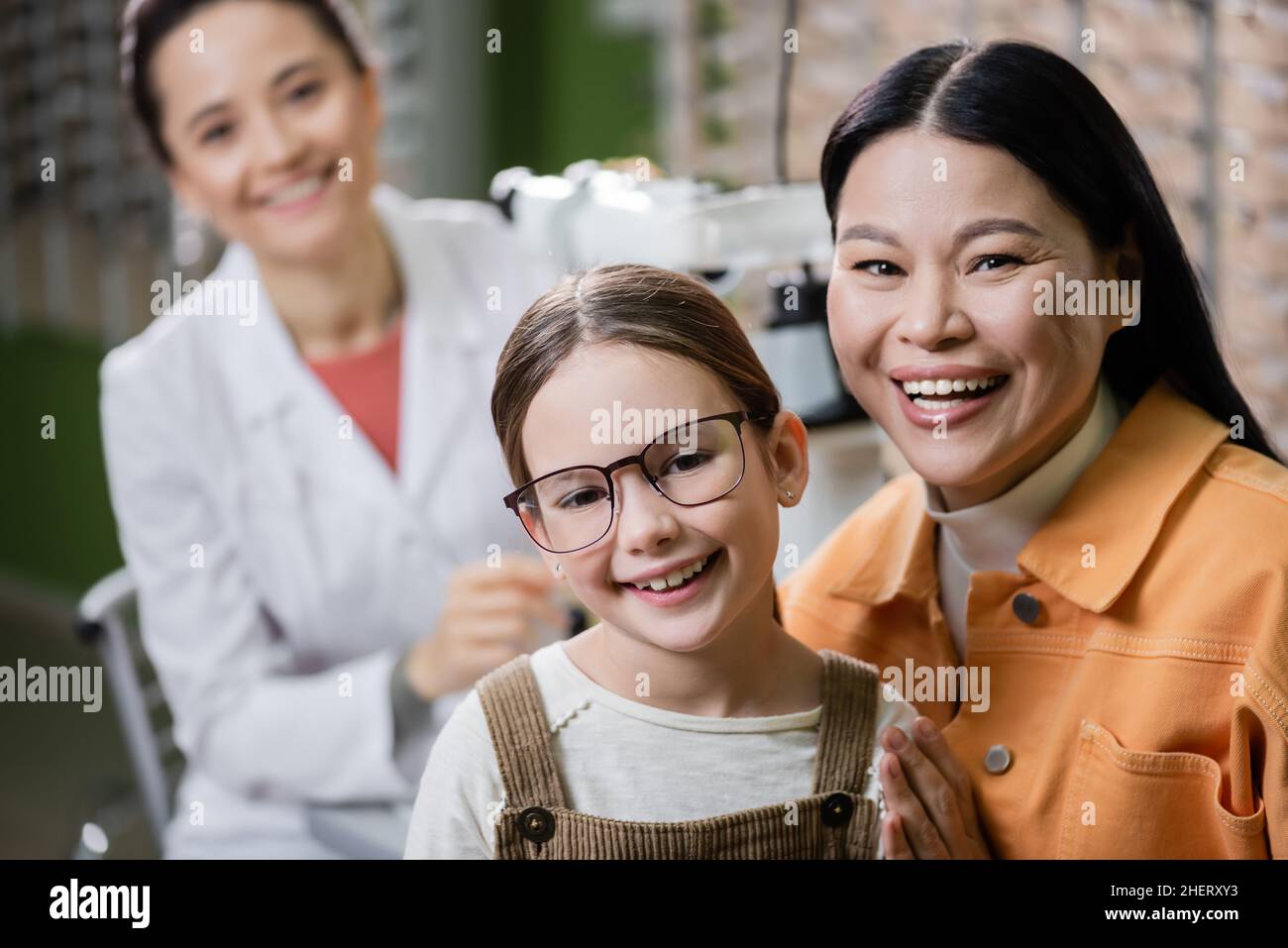 bambino felice in occhiali guardando la macchina fotografica vicino al mom asiatico e oftalmologo sfocato in negozio di ottica Foto Stock